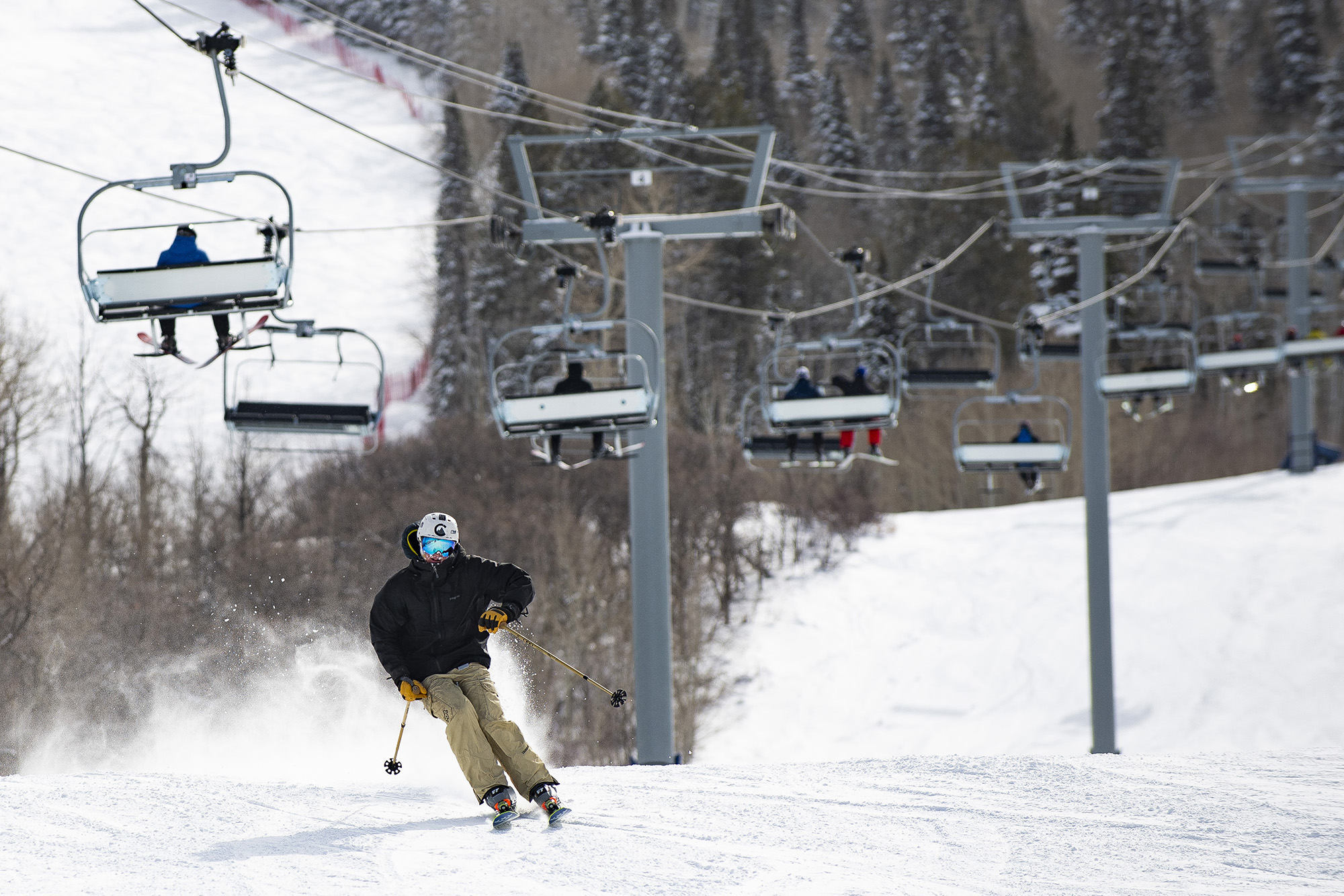 Person skis down snow-covered mountain with several people riding ski lift pictured in background