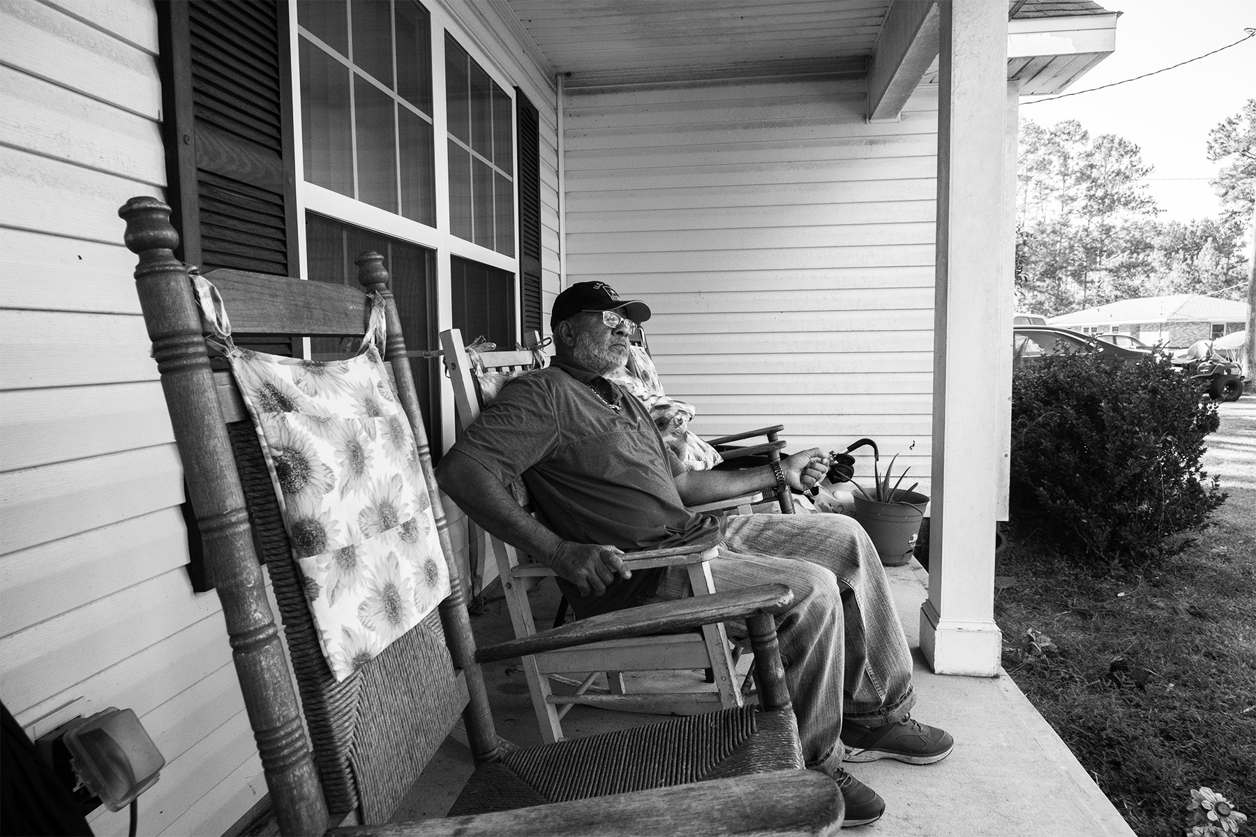 A black and white image of James Siler, wearing a baseball cap and polo shirt, seated in a rocking chair on the front porch of his home in Georgia.