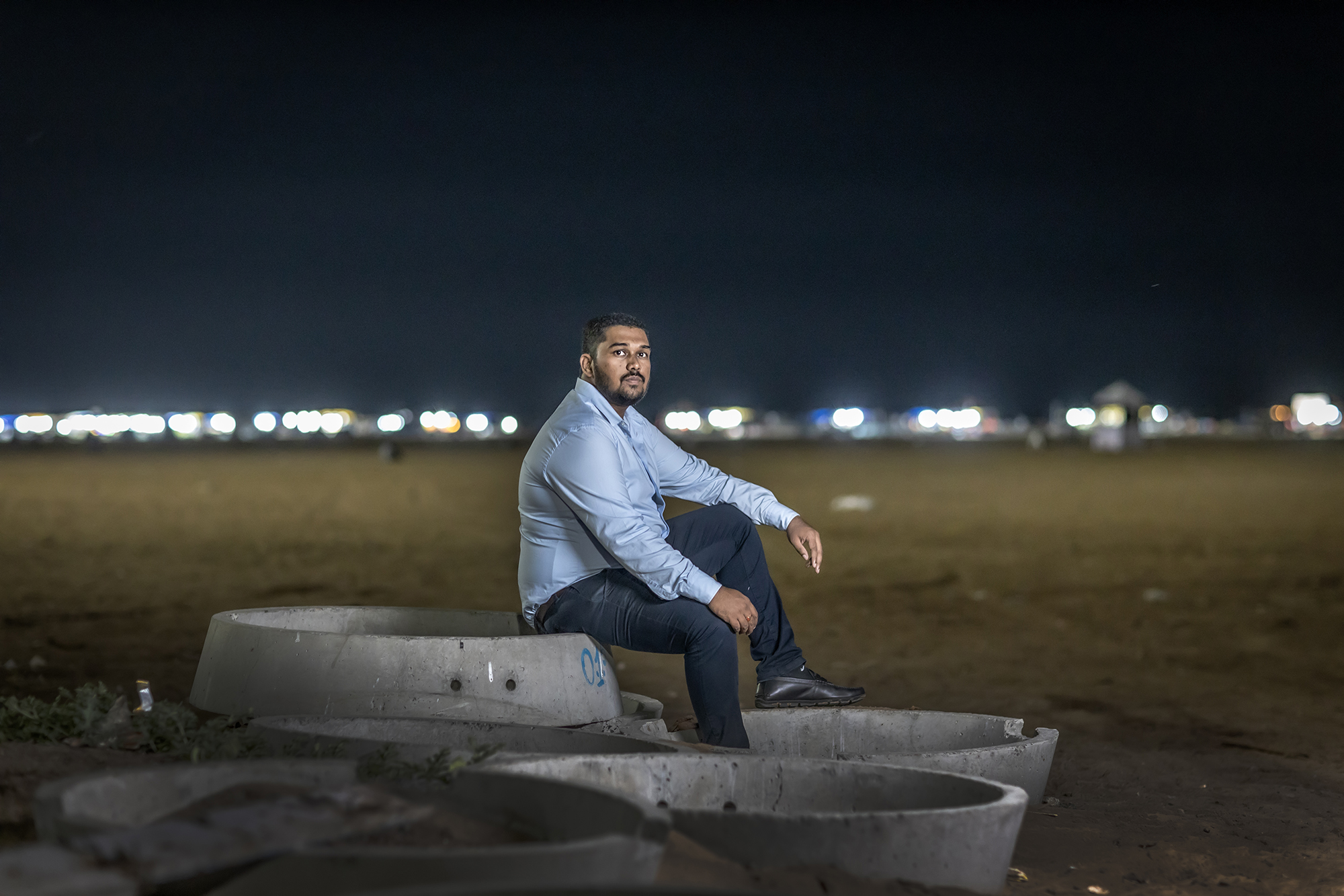 A photo of a man in a dress shirt, slacks and loafers sitting on concrete structures in a field with city lights in the background.