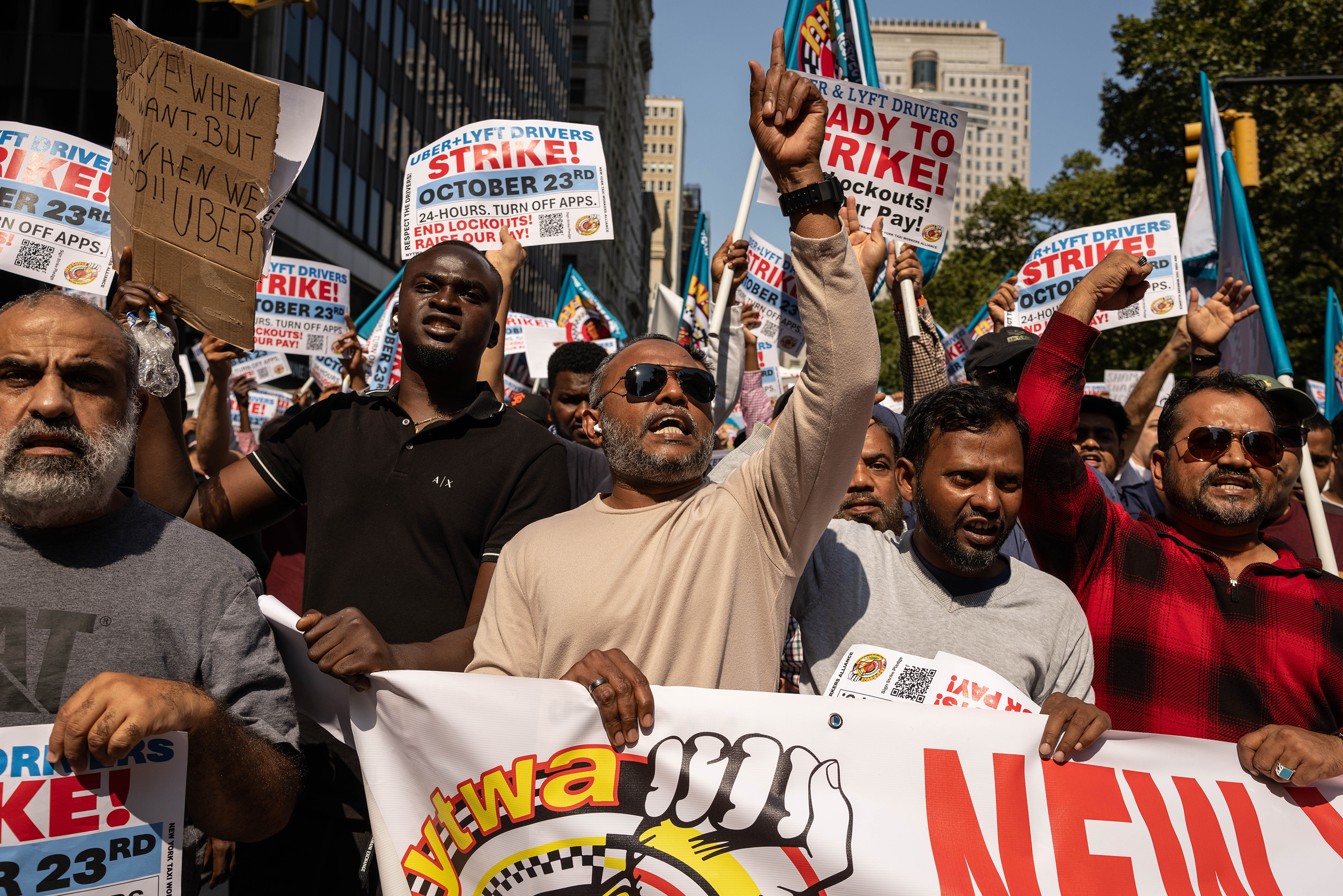A photo showing several people holding signs that say 'Uber+Lyft Drivers Strike,' 'End Lockouts,' and 'Raise Our Pay.'