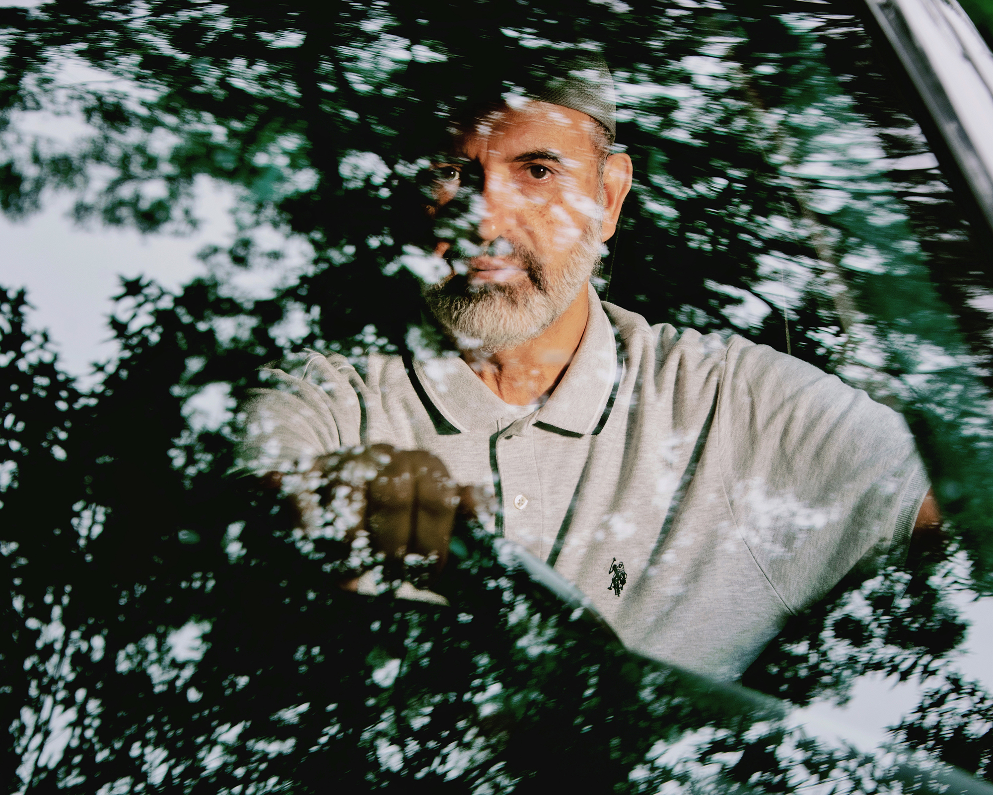 A photo of a man with a white bear and wearing a gray polo in the driver's seat of a car. The windshield of the car has reflections of leaves.