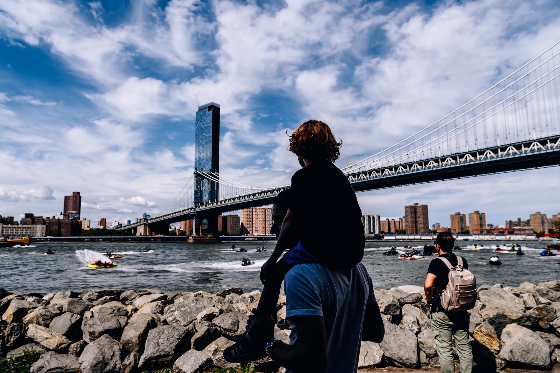 A child on top of an adults shoulders looks out at jet skis in the East River near the Manhattan Bridge.