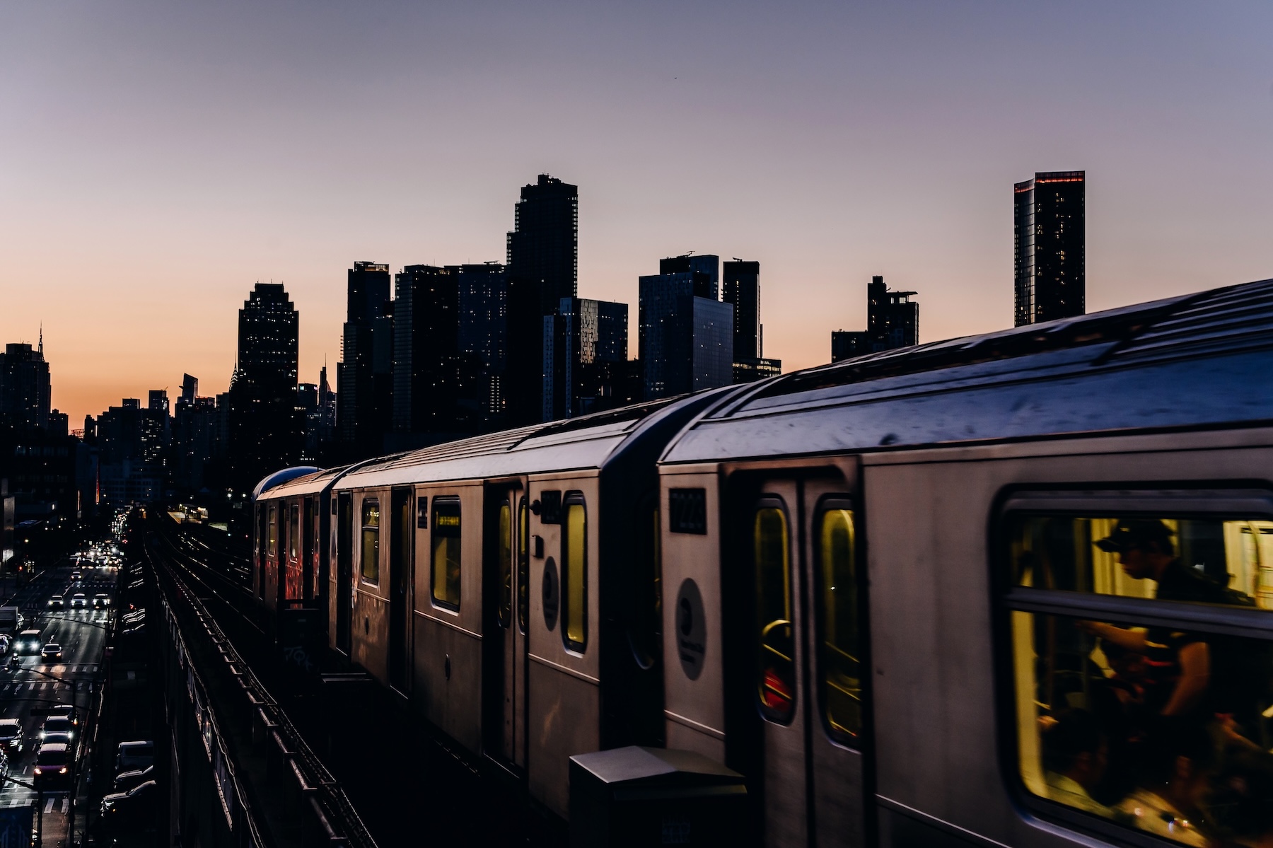A New York subway on an elavated track runs past around sunset. Cars below on the street can be seen, and the New York skyline contrasts a purple and orange sky.