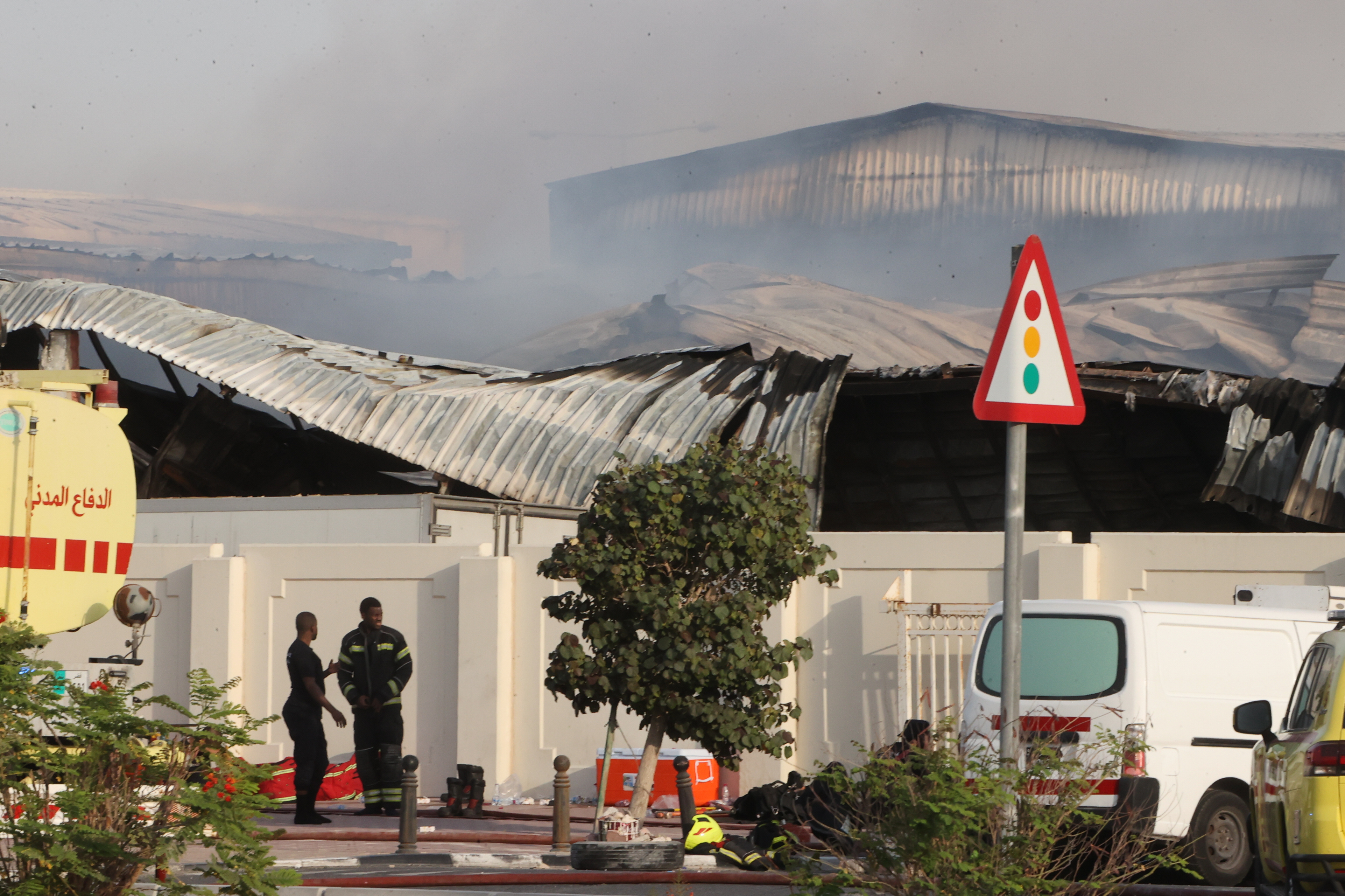 Two people stand outside a building with a collapsed metal roof with smoke risiing from it.