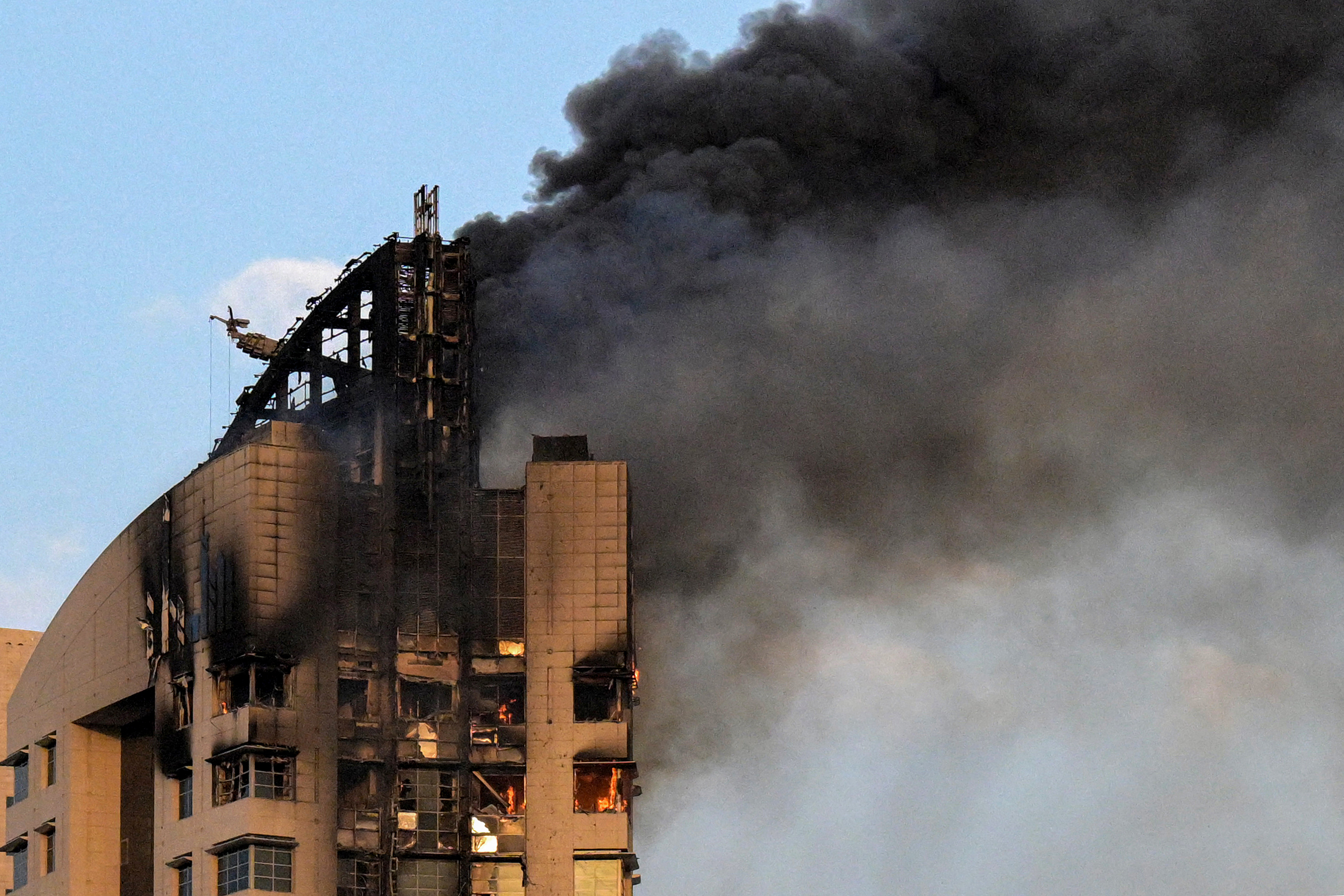 Black smoke billows out of the top of a skyscraper. Flames are visible through windows and holes in the building.